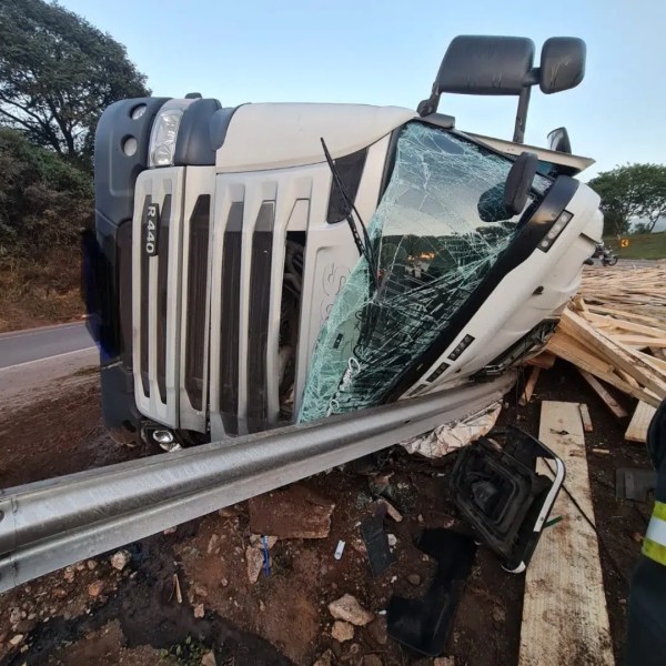 Caminhão R440 tombado na beira da estrada, com o vidro da cabine quebrado e entulho ao redor.