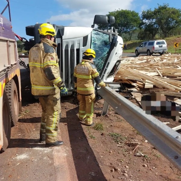 Bombeiros respondendo a um acidente de caminhão tombado na estrada, com carga de madeira espalhada.