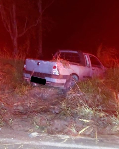 Veículo abandonado em uma área de grama alta à noite, com iluminação vermelha refletindo sobre ele.
