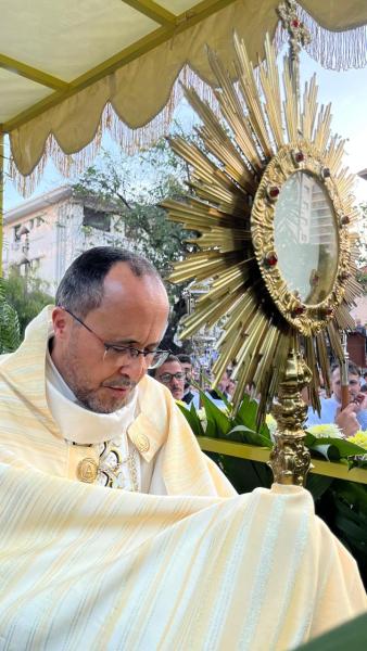 Um sacerdote em um manto claro, em uma cerimônia religiosa, segurando uma custódia dourada com uma hóstia no interior, cercado por foliagem e uma multidão ao fundo.