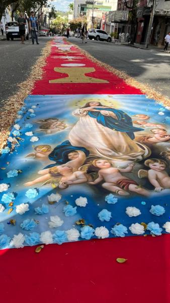 Caminho decorado com tapetes vermelhos e uma grande imagem da Virgem Maria cercada por anjos e flores azuis, em uma rua urbana.