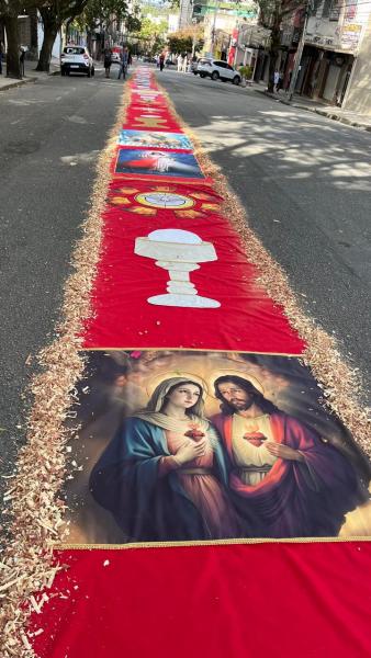 Caminho decorado com tapetes vermelhos, imagens religiosas de Jesus e Maria, e elementos simbolizando a tradição católica, em uma rua urbana.