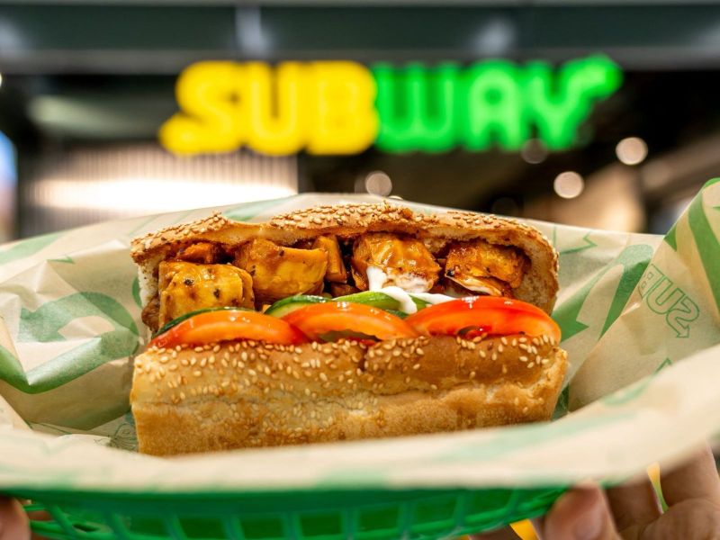 A hand holding a sesame seed sandwich filled with grilled chicken, fresh tomatoes, and green bell peppers, with a Subway sign blurred in the background.