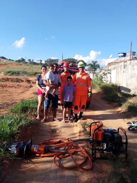 Uma família em pé em uma estrada de terra, com dois bombeiros vestindo uniformes laranjas e capacetes, e um equipamento de resgate ao chão ao lado deles.