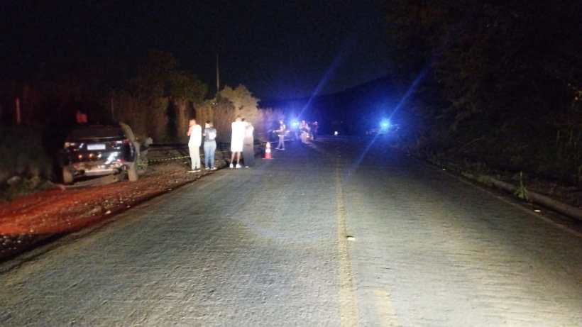 Cena de estrada à noite com pessoas na margem, veículo encostado à direita e luzes de emergência visíveis ao fundo.
