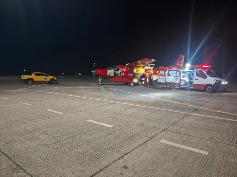 Helicóptero de resgate vermelho estacionado à noite, com uma ambulância ao lado e um veículo amarelo, em um aeroporto.