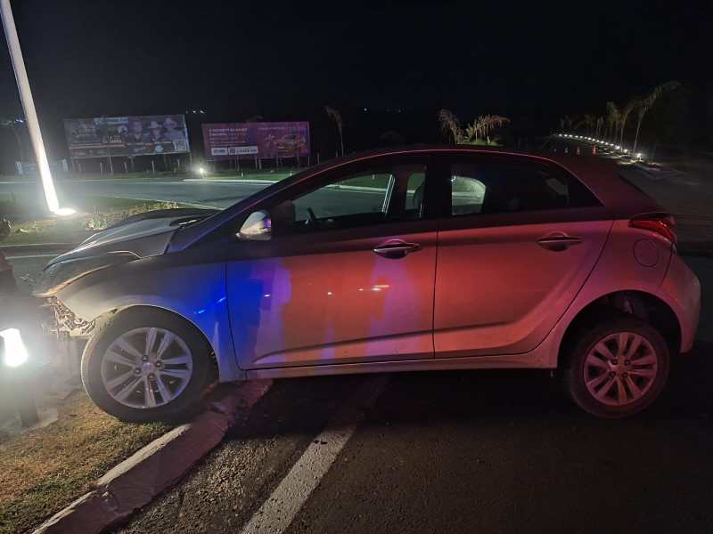 Carro danificado estacionado à noite em uma faixa de pedestres, com luzes refletindo na pintura e outdoors ao fundo.