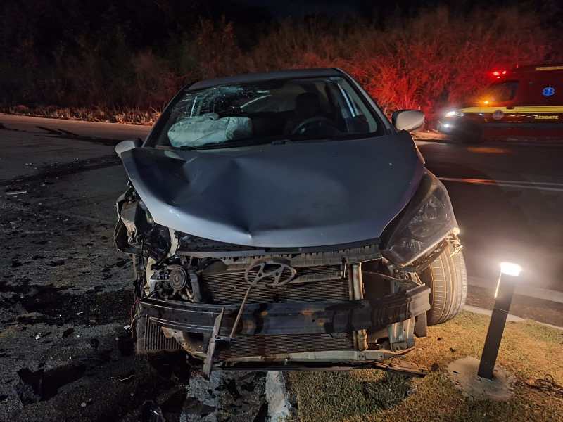 Carro prateado acidentado à noite, com o capô amassado e danos frontais visíveis, ao fundo uma ambulância com luzes de emergência.