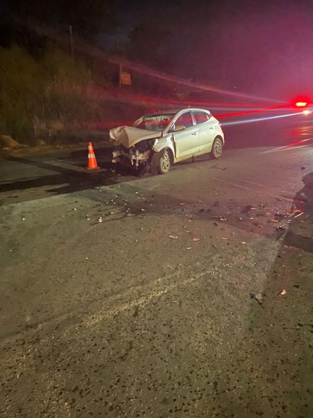 Carro branco danificado na estrada, com cones de sinalização ao redor e debritos espalhados pelo chão.