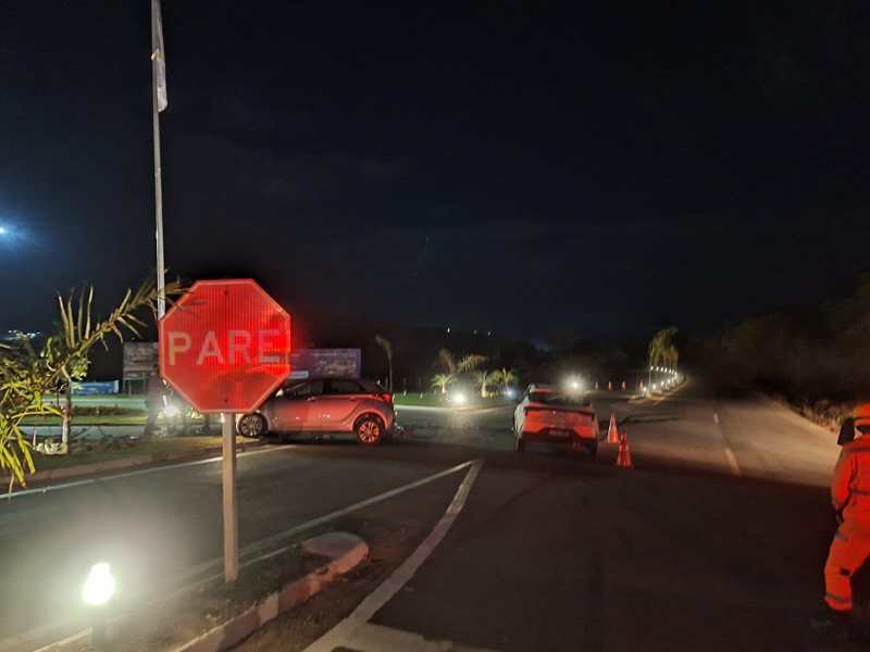Sinal de pare iluminado em uma estrada à noite, com veículos parados e cones de sinalização visíveis.