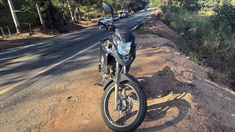 Uma motocicleta preta estacionada ao lado de uma estrada com vegetação e veículos ao fundo.