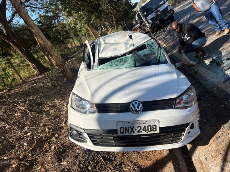 Um carro branco Volkswagen danificado em um acidente, com o teto amassado e o vidro quebrado, parado ao lado da estrada. Dois homens estão próximos, um deles reparando o carro.
