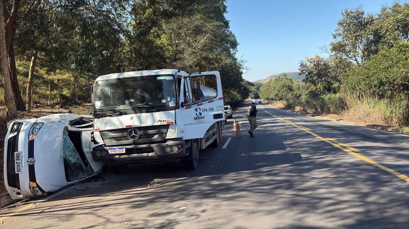Acidente de trânsito com um carro tombado na beira da estrada e um caminhão estacionado ao lado, enquanto uma pessoa inspeciona a cena.