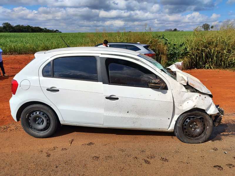 Carro branco danificado com a frente amassada, estacionado em uma estrada de terra ao lado de um campo.