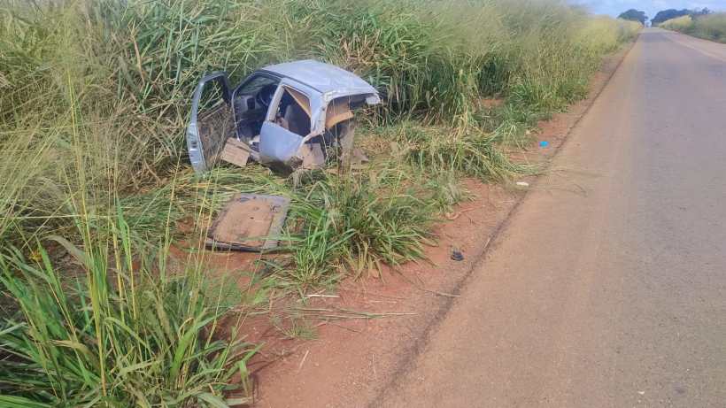 Um carro acidentado parcialmente coberto por vegetação, parado ao lado de uma estrada.