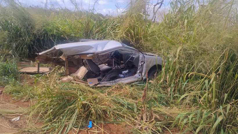 Carro danificado abandonado em meio à vegetação alta.