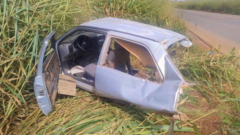 Um carro acidentado abandonado ao lado da estrada, cercado por cana-de-açúcar, com a porta do motorista aberta.