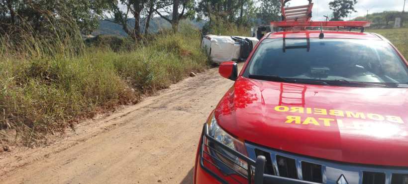 Veículo de emergência dos bombeiros em primeiro plano, com um carro virado ao fundo em uma estrada de terra.