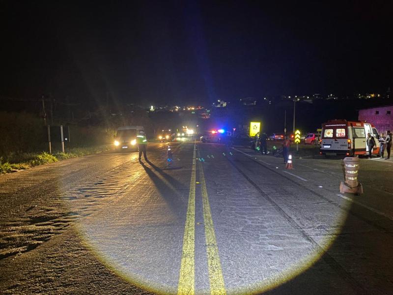 Cena noturna de uma rodovia com veículos parados, incluindo uma ambulância, e presença de pessoas em atividade, com iluminação de emergência visível.