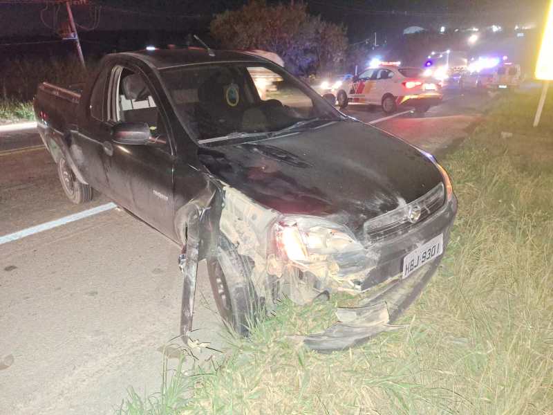 Um carro preto danificado estacionado à beira da estrada à noite, com sinais de um acidente. Patrulhas e luzes de emergência ao fundo.