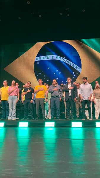 Grupo de pessoas em pé em um palco, cantando, com a bandeira do Brasil ao fundo. Algumas pessoas vestem camisetas amarelas.