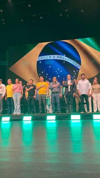 Grupo de pessoas em pé, cantando, com a bandeira do Brasil ao fundo.
