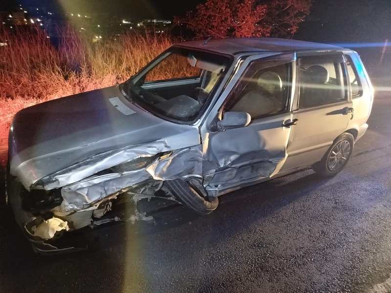 Carro prateado danificado em um acidente, com a lateral amassada e o capô parcialmente aberto, na estrada à noite.