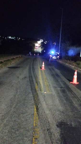 Estrada à noite com marcas de pneu e cones de sinalização, além de um veículo da polícia ao fundo.