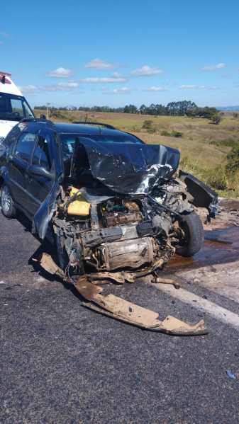 Carro preto destruído em um acidente, com a frente amarga e peças expostas, em uma estrada rural.