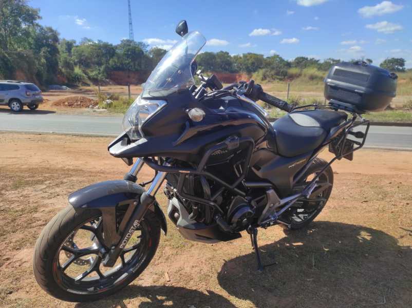 Motocicleta preta em uma estrada com vegetação ao fundo, equipada com bagageiro traseiro.