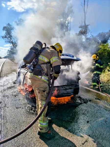 Bombeiros combatendo incêndio em um carro na rodovia com fumaça e chamas visíveis.