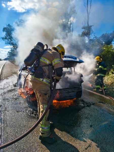 Bombeiros combatendo um incêndio em um carro na estrada, com fumaça e chamas visíveis.