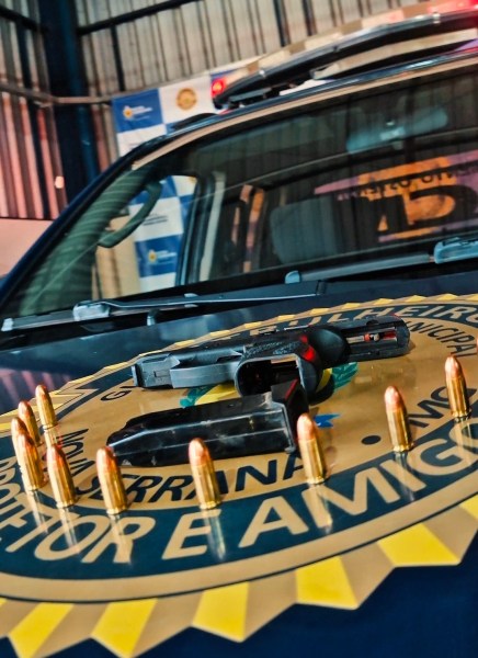 Arma de fogo e munições em cima do capô de um carro policial, com logotipo visível ao fundo.