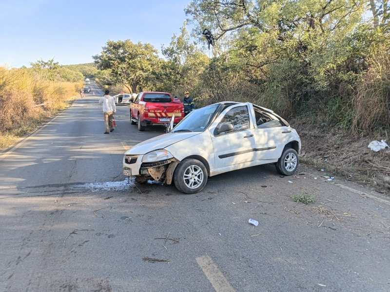 Um carro branco danificado estacionado à beira da estrada, com pessoas ao fundo e vegetação ao redor.