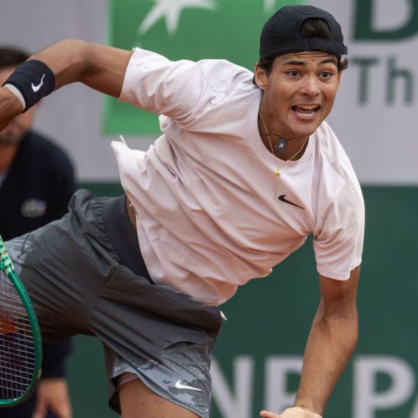 A tennis player in mid-serve, wearing a light pink shirt and gray shorts, showing determination during a match.