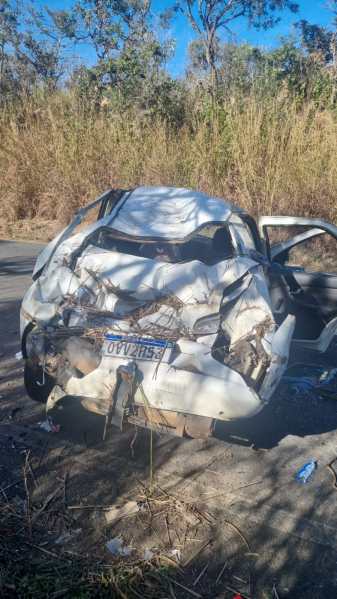 Carro branco capotado e danificado em uma estrada cercada por vegetação seca.