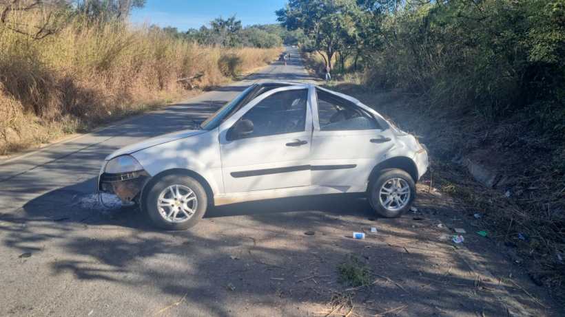 Carro branco parado em uma estrada rural com vegetação ao redor, aparentando danos na parte frontal.