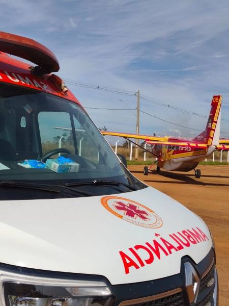 Ambulância de resgate em primeiro plano, com um avião de emergência ao fundo.