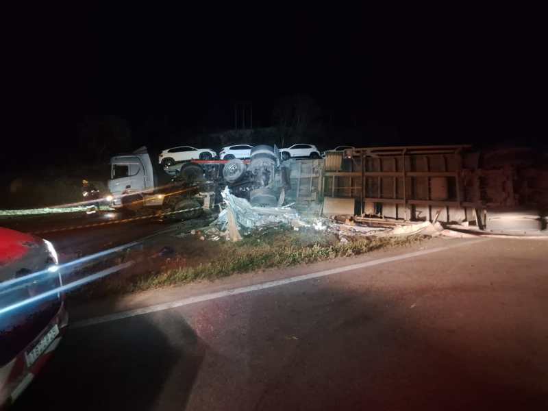 Caminhão tombado na estrada à noite, com outros veículos em cima e uma cena de acidente.