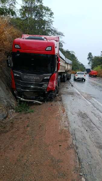 Caminhão Scania vermelho parcialmente saindo da estrada em uma zona rural, com um veículo branco passando ao fundo e vegetação ao redor.