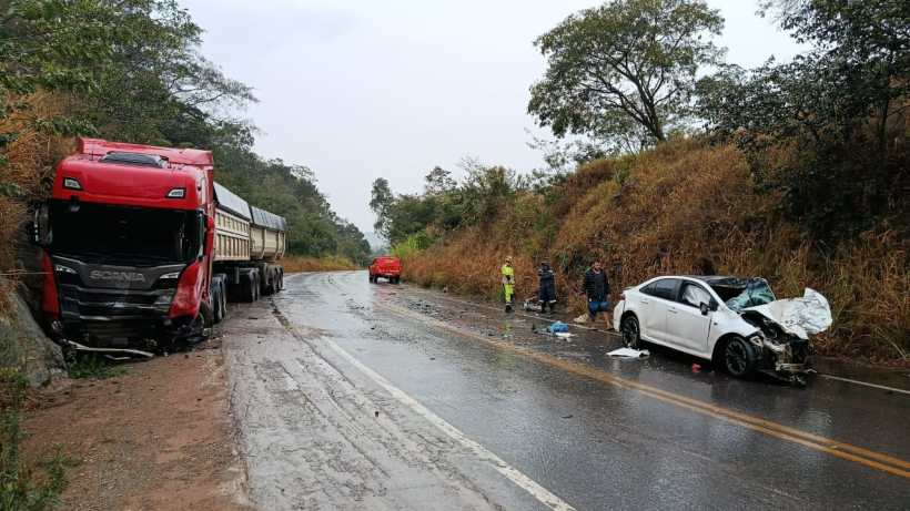 Acidente de trânsito em uma estrada, com caminhão vermelho tombado, carro branco danificado e pessoas assistindo no local.