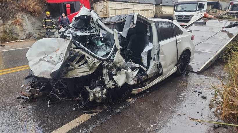 Carro destruído após acidente na estrada, com danos significativos na lateral e na frente, e bombeiros e pessoas ao fundo.