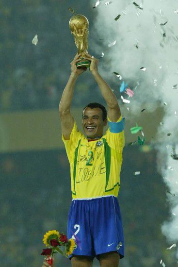 A Brazilian football player celebrates by holding the FIFA World Cup trophy high above his head, surrounded by confetti and smoke.