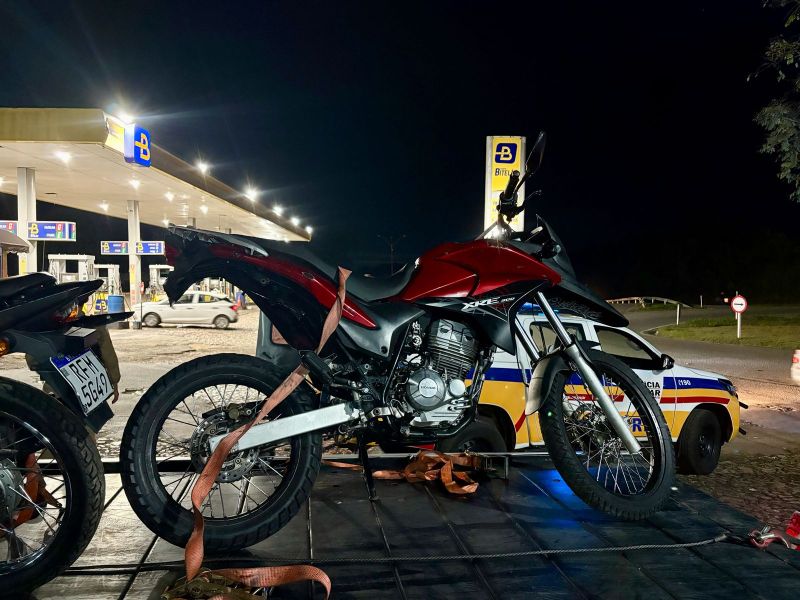 Duas motocicletas sendo transportadas em um veículo, com uma delas em destaque, de cor vermelha, estacionadas em uma área próxima a um posto de gasolina à noite.