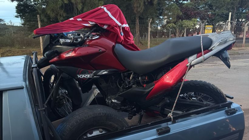 Moto vermelha sendo transportada em uma caminhonete, coberta com uma lona. Pneus visíveis na caçamba.