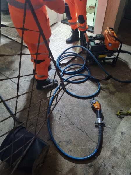 Pessoas em macacões laranja operando um equipamento com mangueiras em um ambiente de trabalho, com uma estrutura de ferro ao fundo.