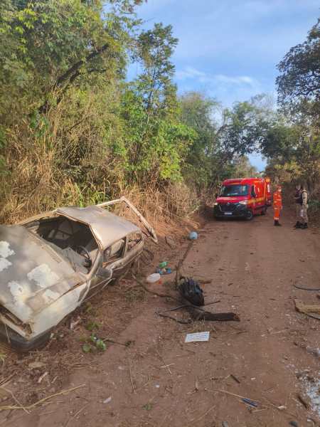 Carro abandonado em estado deteriorado ao lado de uma estrada de terra, com um veículo de resgate e várias pessoas no fundo cercados por vegetação.