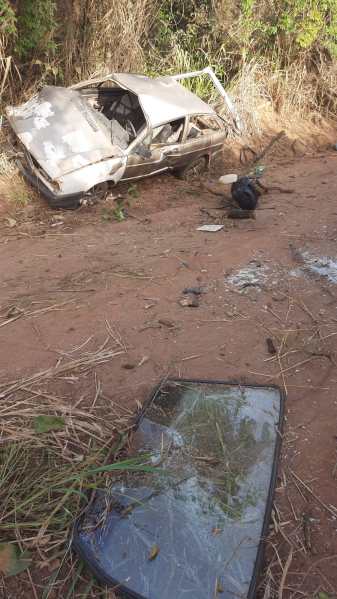 Um carro velho abandonado em um terreno, cercado por vegetação. Há um vidro quebrado próximo ao veículo.