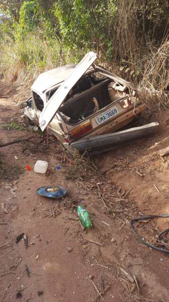 Um carro abandonado e danificado em um terreno acidentado, com a capota levantada e do lado de uma estrada de terra. Objetos como uma garrafa plástica e utensílios estão espalhados ao redor.