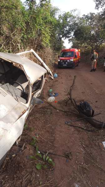 Veículo acidentado em uma estrada rural, com um caminhão de emergência ao fundo e um agente de resgate ao lado.
